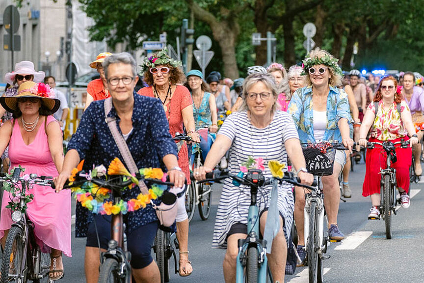 Bunt gekleidete Frauen auf geschmückten Fahrrädern lachend auf einer Straße radelndf ei